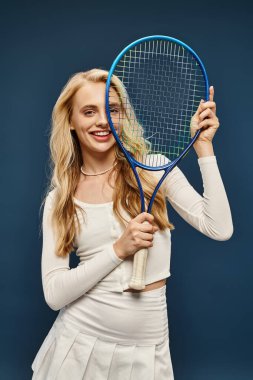 Tenis raketiyle beyaz şık giyinmiş heyecanlı sarışın kadın mavi kameraya bakıyor.