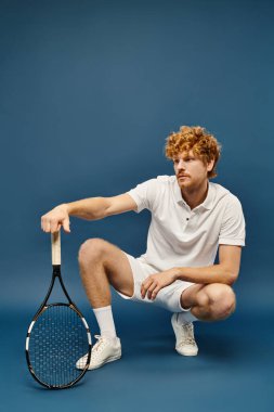 trendy redhead man in white tennis outfit sitting on haunches with racquet on blue, old money style clipart