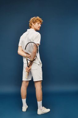 Beyaz polo tişörtlü ve şortlu kızıl saçlı adamın arkadan görünüşü mavi tenis raketiyle duruyor.