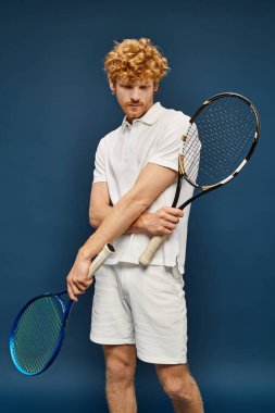 aristocratic redhead man in white outfit posing with tennis racquets posing on blue backdrop clipart