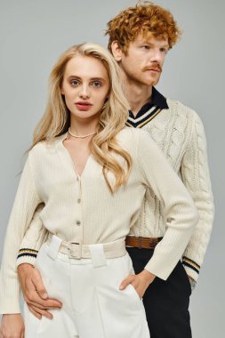 Gri arka planda şık giyinmiş kızıl saçlı adamın yanındaki kameraya bakan çekici sarışın kadın.