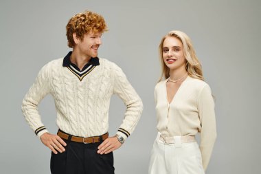 Elleri kalçasında neşeli kızıl saçlı adam gri elbiseli, modaya uygun sarışın kadına bakıyor.