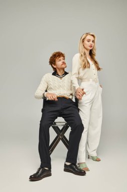 Gri arka planda oturan ve moda kızıl saçlı adamın elini tutan çekici sarışın kadın.