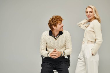 Şık, kızıl saçlı bir adam, sarışın bir kadının yanında oturuyor. Gri arka planda kameraya gülümsüyor.