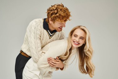 cheerful redhead man hugging excited blonde woman and having fun on grey backdrop, classic fashion clipart