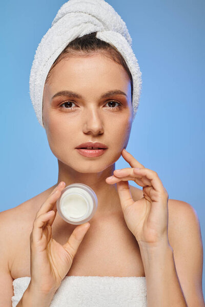 portrait of woman with towel on head holding jar of face cream on blue backdrop, wellness and beauty