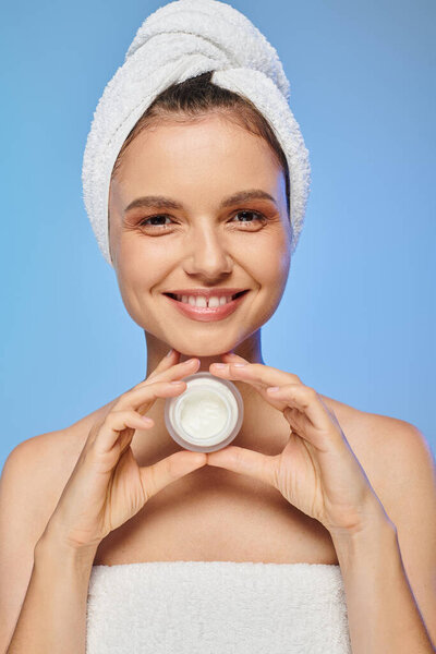 joyful woman with towel on head holding jar of face cream on blue backdrop, wellness and beauty