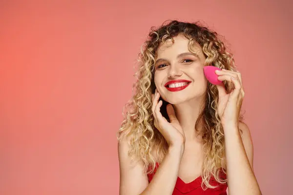 stock image curly joyful model with red lips using face sponge on pink and yellow backdrop, elegant beauty