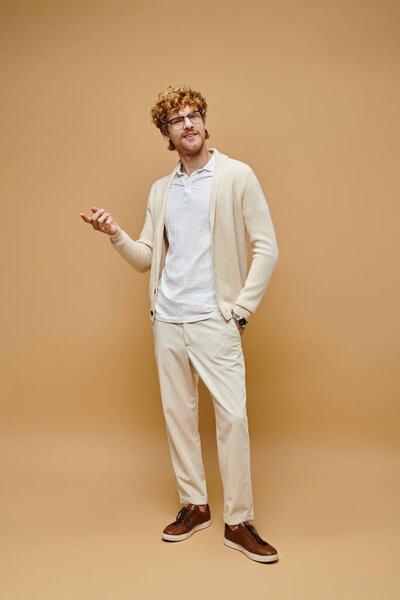 Полная длина элегантный рыжий мужчина в очках и модной одежде с рукой в кармане на бежевый