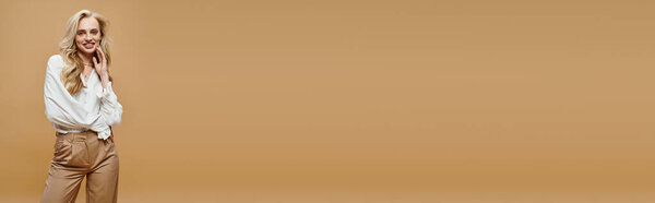 веселая блондинка в стильной блузке и брюках, улыбающаяся в камеру на бежевом фоне, баннер
