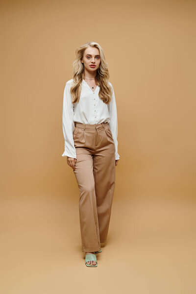 блондинка полной длины в белой блузке и брюках смотрит на камеру в бежевом стиле старых денег