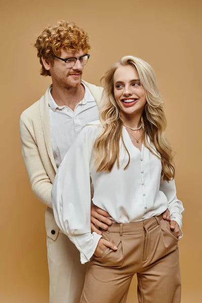 stock image redhead man in eyeglasses embracing blonde woman standing with hands in pockets on beige, fashion