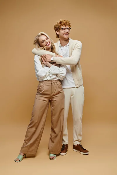 stock image full length of smiling redhead man in eyeglasses hugging blonde woman on beige, old money style