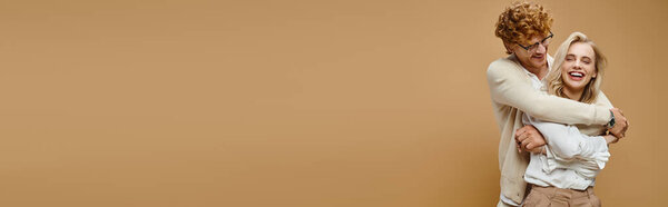 рыжеволосый мужчина в очках обнимает смеющуюся блондинку на бежевом, горизонтальном баннере