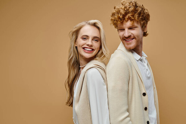 joyous young models in light-colored vintage attire standing back to back on beige backdrop