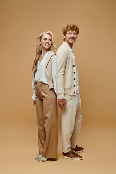 full length of trendy couple in old money style clothes holding hands standing back to back on beige