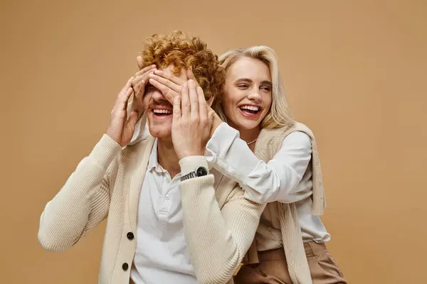 stock image playful blonde woman covering eyes of stylish redhead man on beige, old money style fashion