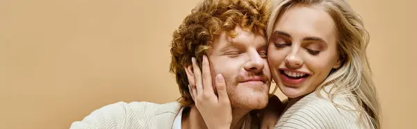 stock image overjoyed blonde woman with closed eyes embracing head of trendy redhead man on beige, banner