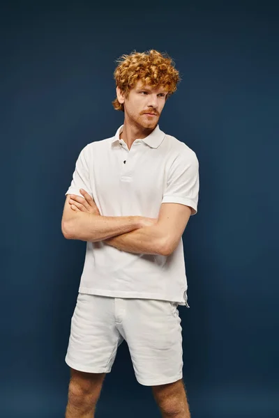 stock image redhead young man in white polo t-shirt and shorts posing with folded arms on blue backdrop