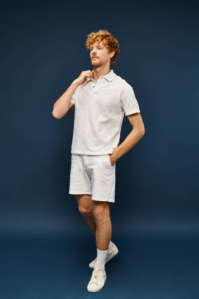 redhead young man in white polo t-shirt and shorts standing with hand in pocket on blue backdrop