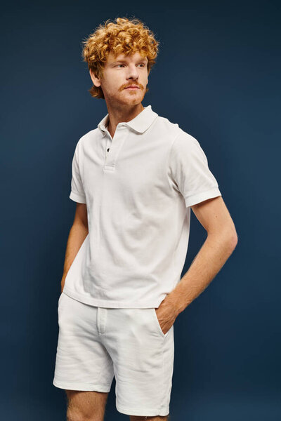 redhead man in white polo t-shirt and shorts posing with hands in pockets and looking away on blue
