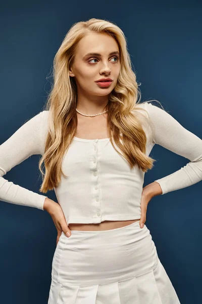 stock image attractive blonde woman in white stylish  attire standing with hands on hips looking away on blue