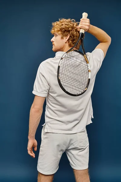 stock image back view of young redhead man in white tennis outfit posing with racquet on blue, timeless fashion