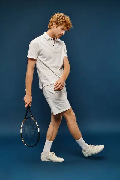 stock image full length of young redhead man in white trendy outfit walking with tennis racquet on blue backdrop