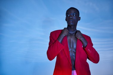 Pembe pantolonlu ve kırmızı ceketli şık, Afro-Amerikan bir adam sulu arka plana bakıyor.