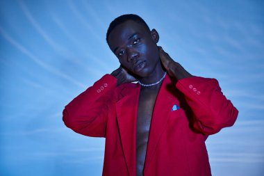 Kırmızı renkli ceketli, mavi sulu arka planda poz veren Afro-Amerikan bir adam.