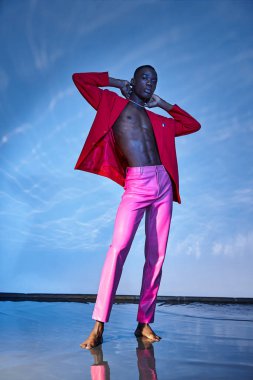 Baştan çıkarıcı Afro-Amerikan erkeği pembe pantolonlu ve kırmızı ceketli mavi arkaplanda poz veriyor.