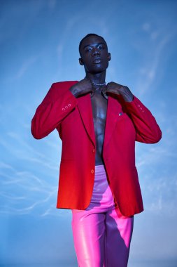 Pembe pantolonlu, kırmızı ceketli ve mavi arkaplanda poz veren afro-amerikalı çekici bir adam.