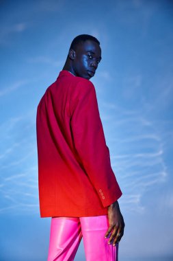 Pembe pantolonlu, kırmızı ceketli ve mavi arkaplanda poz veren afro-amerikalı çekici bir adam.