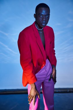 Pembe pantolonlu, kırmızı ceketli ve mavi arkaplanda poz veren Afro-Amerikan yakışıklı adam.