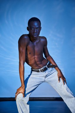 Şık kotlu, gömleksiz Afro-Amerikan bir adam. Arka plandaki kameraya bakıyor.