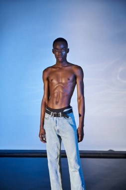 Gömleksiz, stil sahibi, modern kot pantolonlu ve mavi arka planda kameraya bakan Afro-Amerikan bir adam.