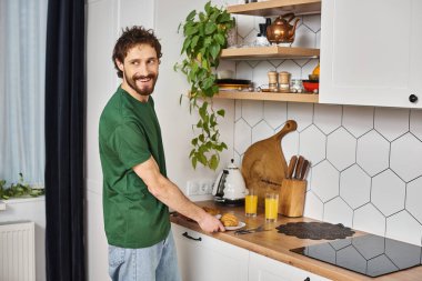 handsome cheerful man in casual comfy attire preparing to eat croissants and looking away in kitchen clipart