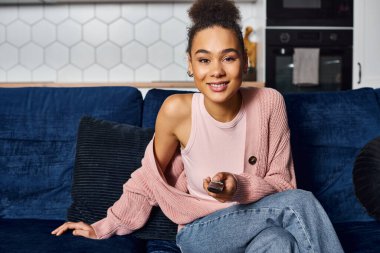 Güzel Afrikalı Amerikalı neşeli kadın kanepeye oturmuş, uzaktan kumandayla kameraya bakıyor.