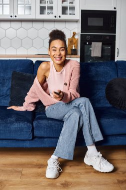 Güzel, neşeli, Afro-Amerikalı kadın, günlük giysiler içinde, kanepede oturuyor ve evde televizyon seyrediyor.