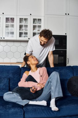 Güzel, neşeli, Afro-Amerikalı kadın kanepeye oturmuş ve evdeki neşeli erkek arkadaşına bakıyor.