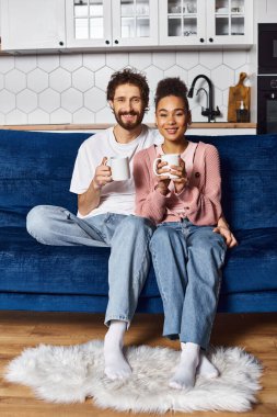 Ev elbiseli neşeli, çok kültürlü bir çift kanepede kahve içip kameraya gülümsüyor.