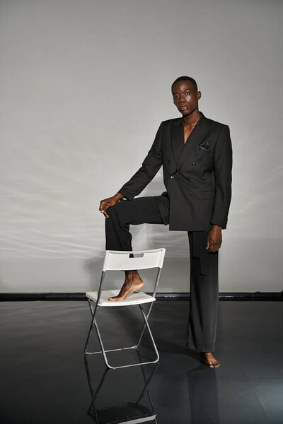tempting african american man in sexy black suit posing next to chair on gray watery backdrop