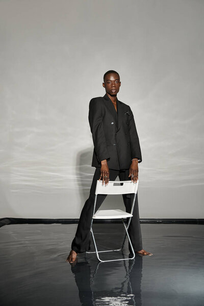 charming african american man in sexy black suit posing next to chair on gray watery backdrop