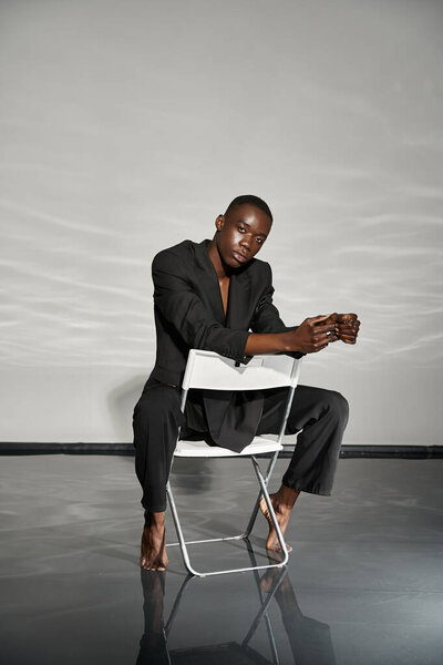 appealing sexy african american man in elegant black suit sitting on chair and looking at camera