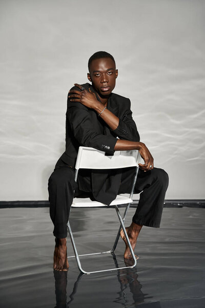 appealing sexy african american man in elegant black suit sitting on chair and looking at camera