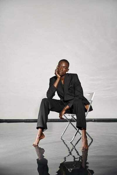 trendy sexy african american man in elegant black suit sitting on chair and looking at camera