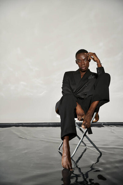 stylish sexy african american man in elegant black suit sitting on chair and looking at camera