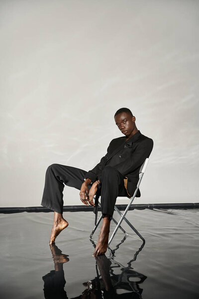 stylish sexy african american man in elegant black suit sitting on chair and looking at camera
