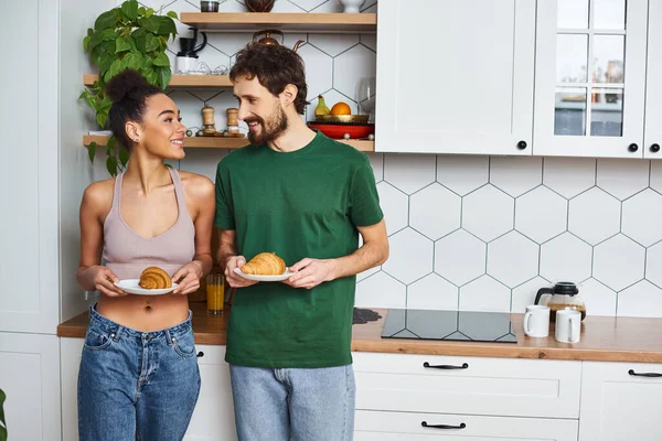 Stock image diverse joyous couple in cozy homewear holding plates with delicious croissants while in kitchen