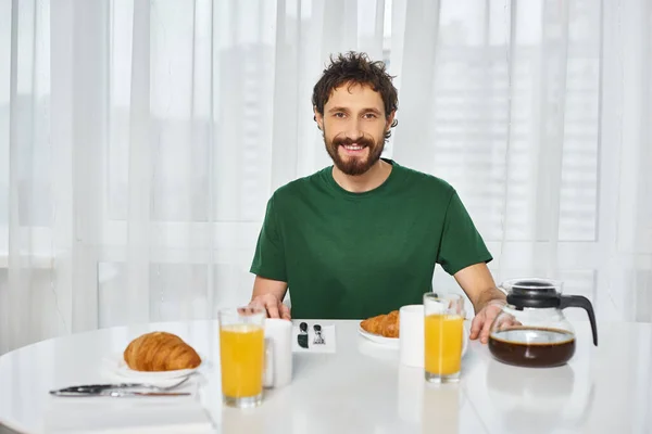 cheerful attractive man in casual attire sitting at table during breakfast and smiling at camera